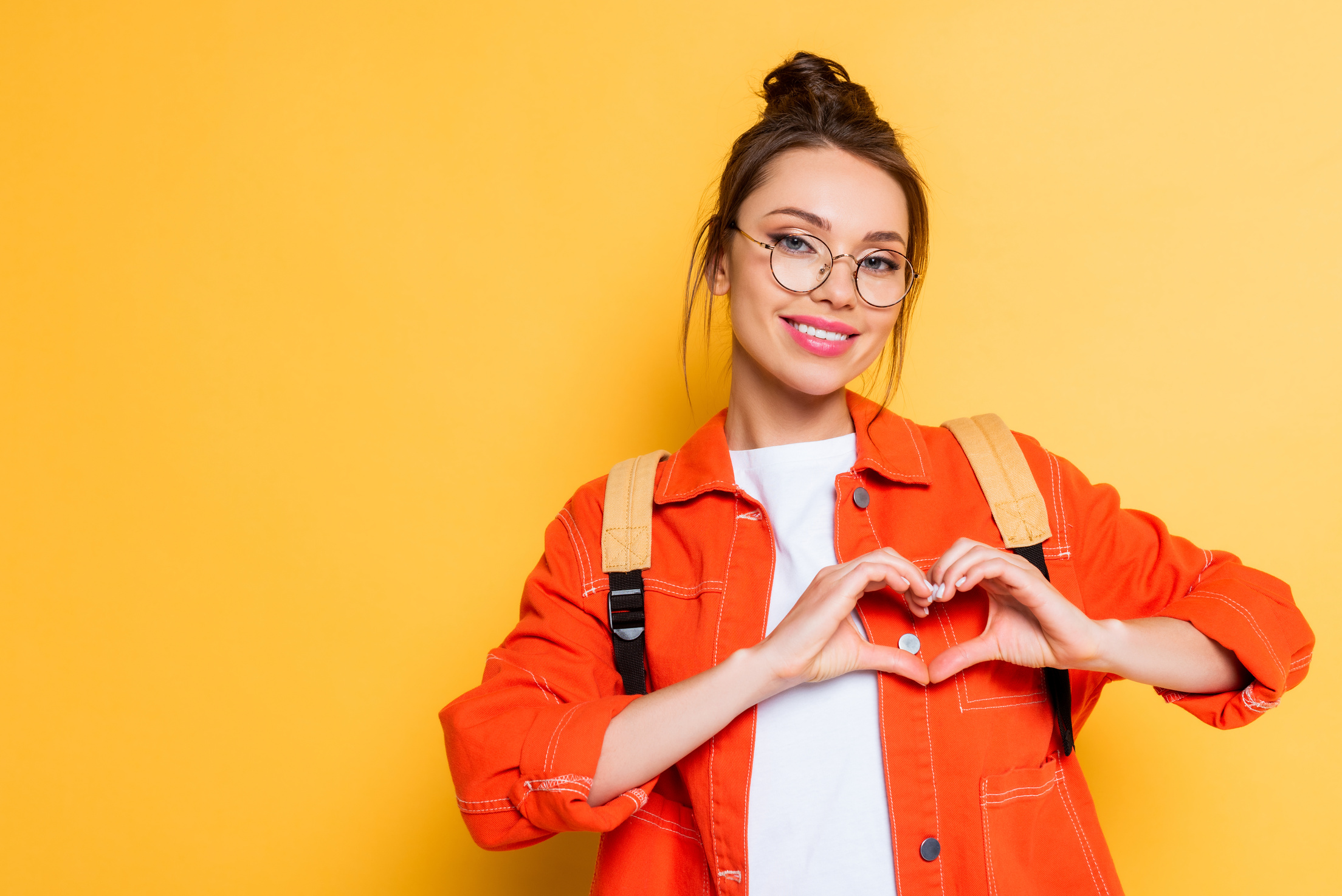 happy student in eyeglasses showing heart sign with hands on yellow background